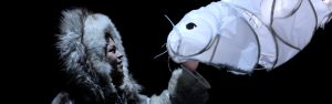 Actor dressed in fur parka looks fondly at a paper machier seal while on stage in a production of Stories That Transform Us.